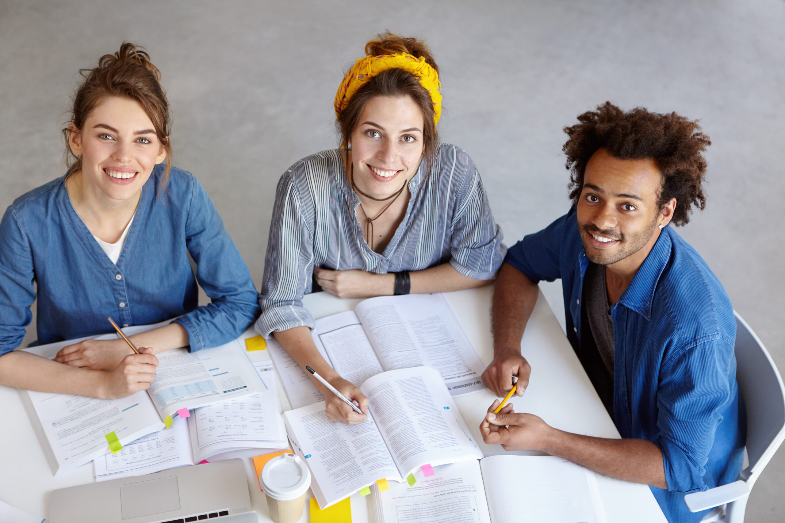 International friendship and cooperation concept. Two females helping their African dark-skinned friend male with studying showing him information in books, explaining him new material and hard theory
